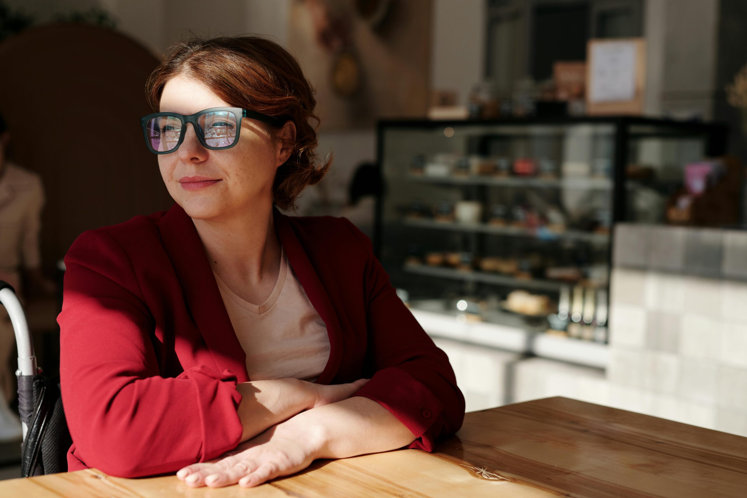 Woman in a wheelchair enjoying a sunny day at a café, wearing eyeglasses and a red jacket.